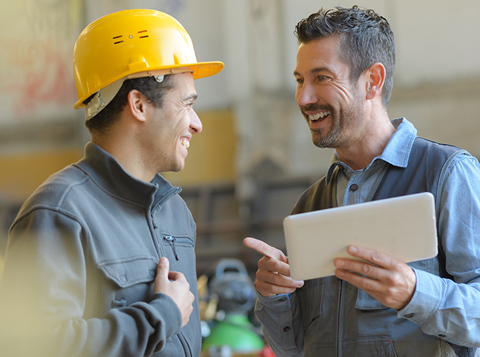 Smiling man holding a tablet talking to another smiling man wearing a hard hat in a warehouse