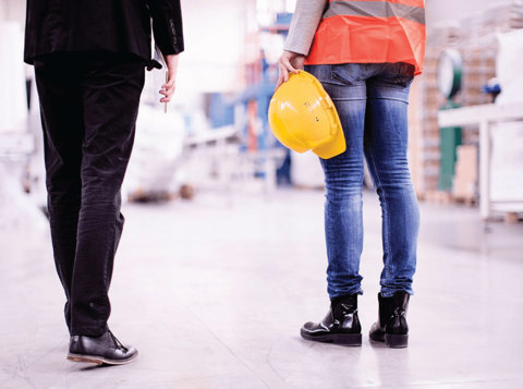 Person in suit walking next to person in high vis holding hard hat in a warehouse