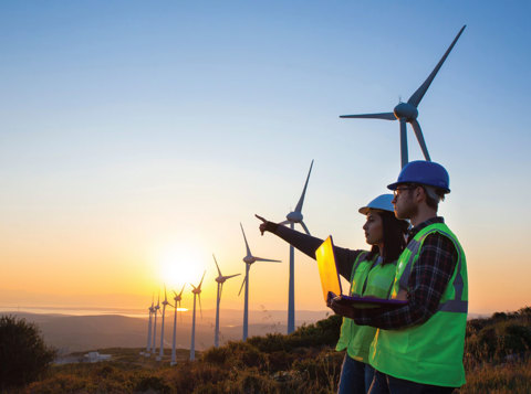 Two people wearing high vis and PPE stood in a wind turbine field holding a laptop and pointing to the distance